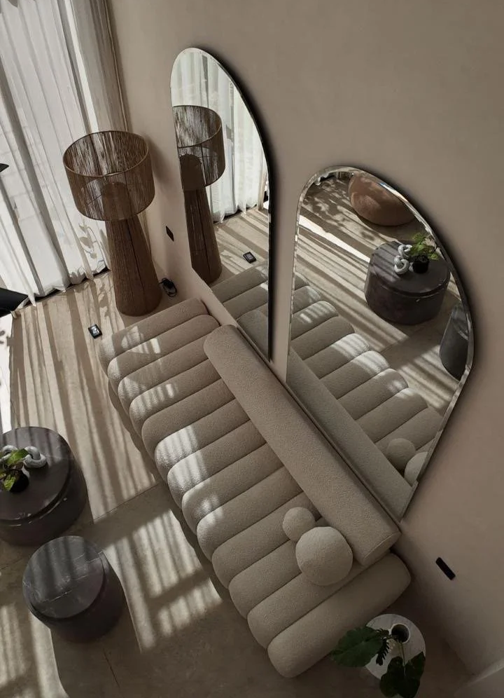 Living room with beige tufted sofa, large curvy mirrors on wall, two tall brown side tables with lamps, round black tables, potted plants, and sunlight casting shadows through curtains.