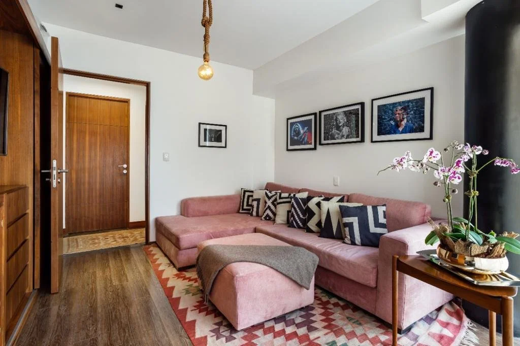 Living room with a pink velvet sectional sofa, patterned pillows, a wooden side table with potted orchids, framed photographs on the wall, wood flooring, and a wooden door.