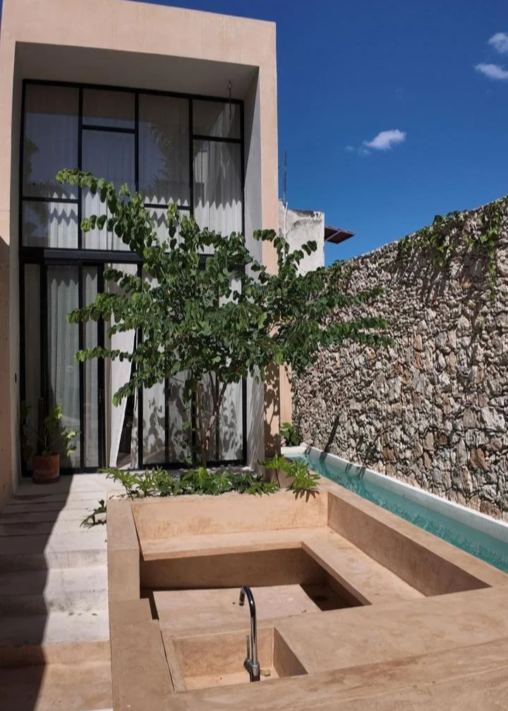 A modern outdoor space with a built-in stone hot tub, a small pool, a large leafy tree, a stone wall, and a two-story glass window house under a blue sky.
