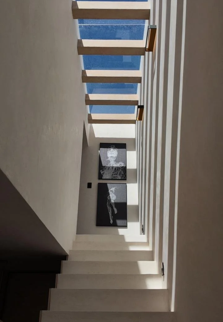 Interior view of a modern staircase with wooden steps and vertical railings, leading upward to a skylight and artwork on the wall below.