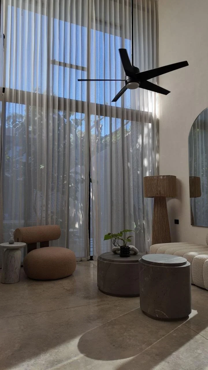 Living room with large windows covered by sheer white curtains, ceiling fan, beige and wooden furniture, green potted plant on marble coffee table, and neutral-colored decor.