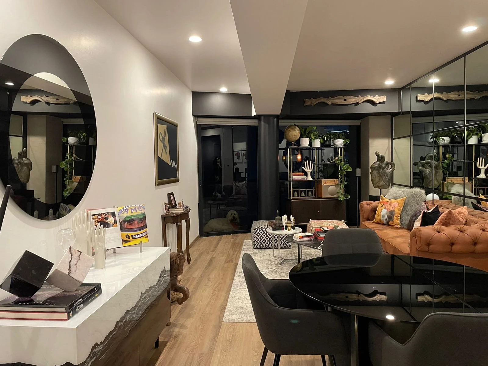 Modern living room with a black round dining table, black chairs, a tan tufted sofa with colorful pillows, a mirrored wall, and decorative wall art and sculptures.