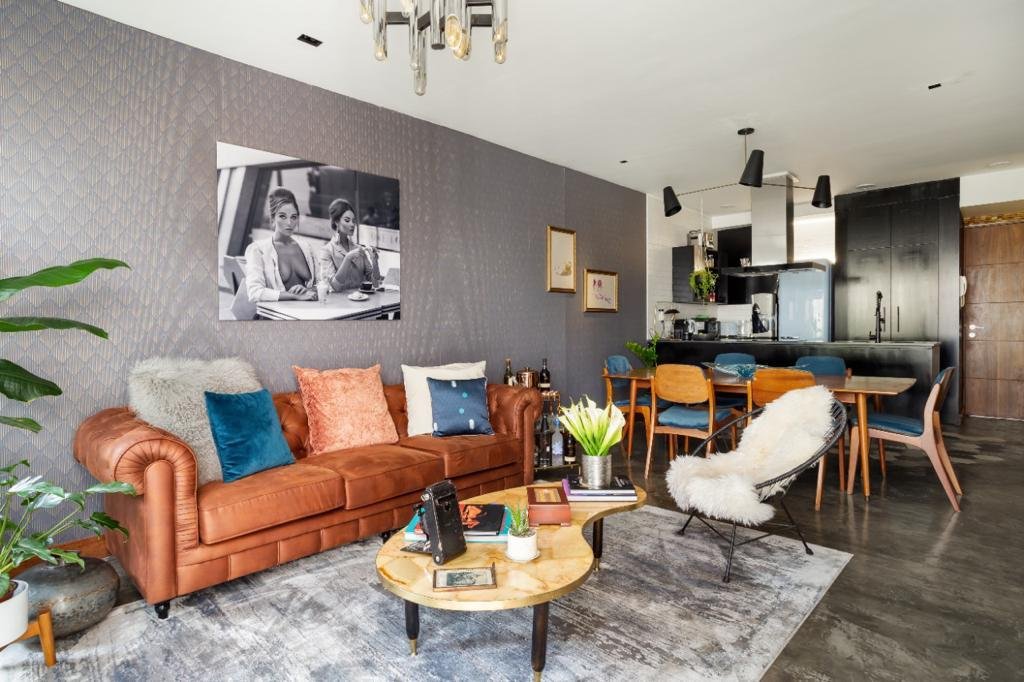 Modern open-concept living room and kitchen with mid-century furniture, including a leather sofa, colorful pillows, and a sheepskin chair. Decorative wall art and a large potted plant are also visible, with the kitchen featuring black cabinetry and a