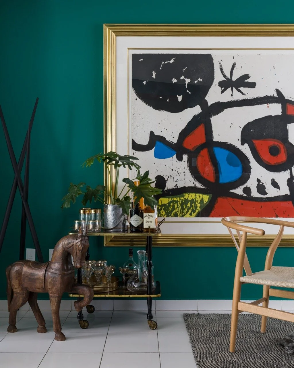 Interior of a room with green wall, large abstract colorful artwork in a gold frame, a wooden horse figurine, a bar cart with plants, bottles, and glasses, a woven chair, and a modern sculpture on the floor.