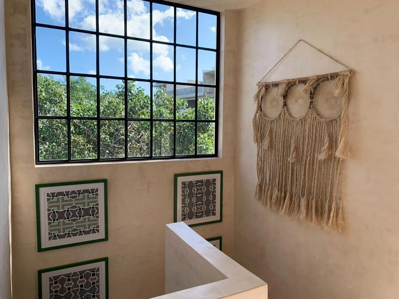 Indoor staircase landing with a large grid window showing trees and a blue sky outside, decorated with two geometric framed artworks on the wall and a large woven wall hanging.
