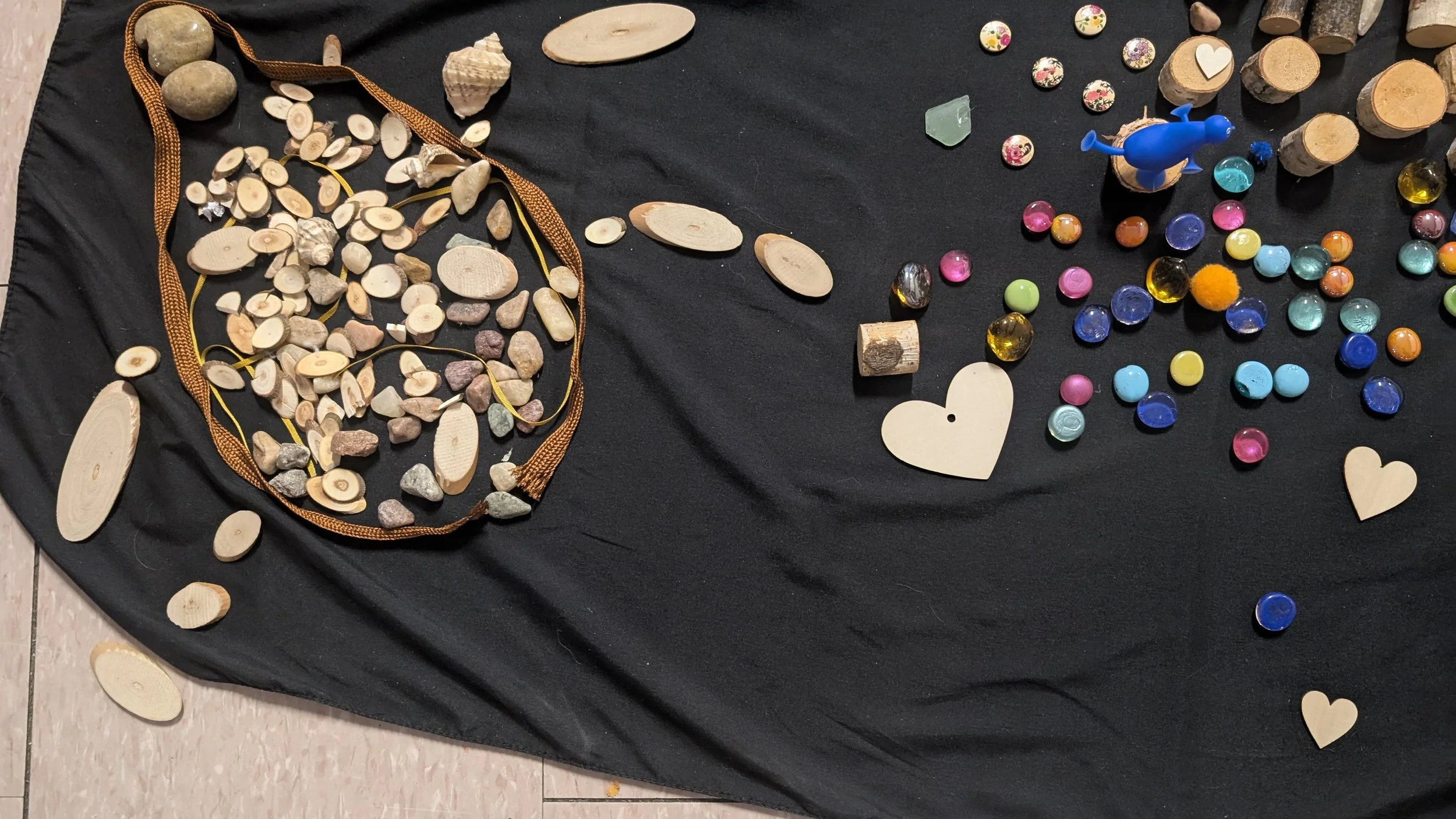 A black cloth covered surface with various craft supplies including wooden hearts, small stones, colorful glass pebbles, some cork pieces, a blue plastic figurine, and small decorative buttons with floral patterns.
