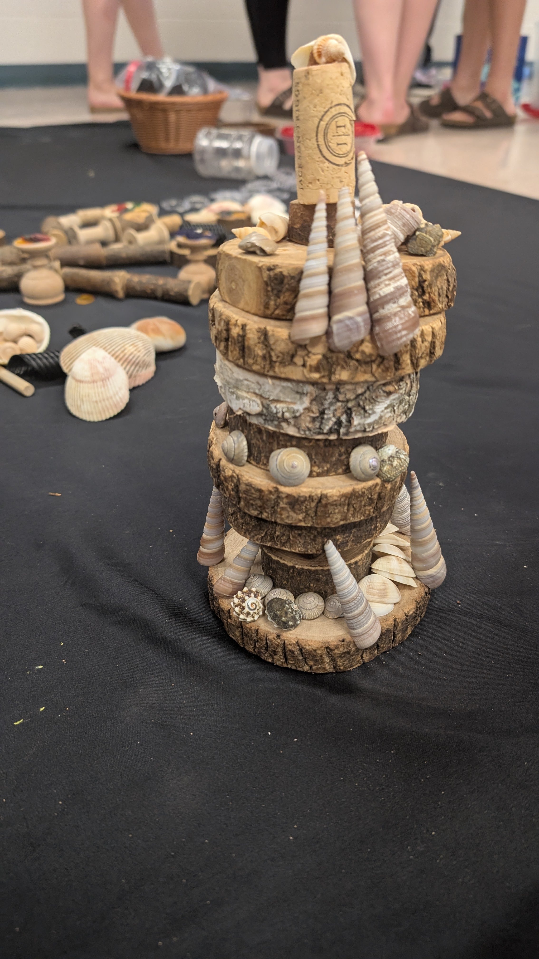 A small sculpture made of stacked wooden disks, shells, and small rocks, arranged to resemble a lighthouse with seashells and rocks around the base, on a black table. In the background, there are blurred people and various objects.