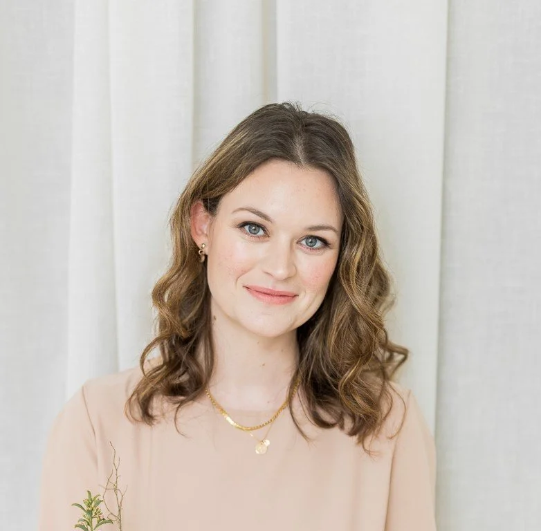 A woman with shoulder-length wavy brown hair and light skin, smiling at the camera, wearing a light pink blouse and gold jewelry, standing in front of a white curtain.