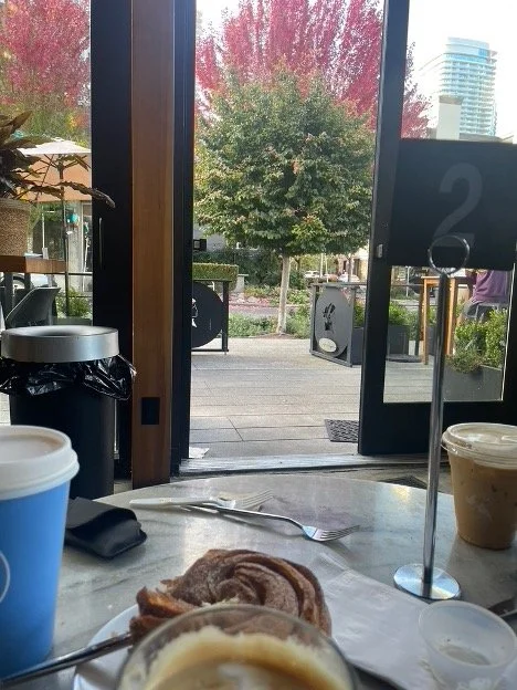 Indoor cafe table with a fork, a napkin, and a cinnamon roll, with a large coffee cup and a dessert container in the background, overlooking an outdoor patio with trees and city buildings.