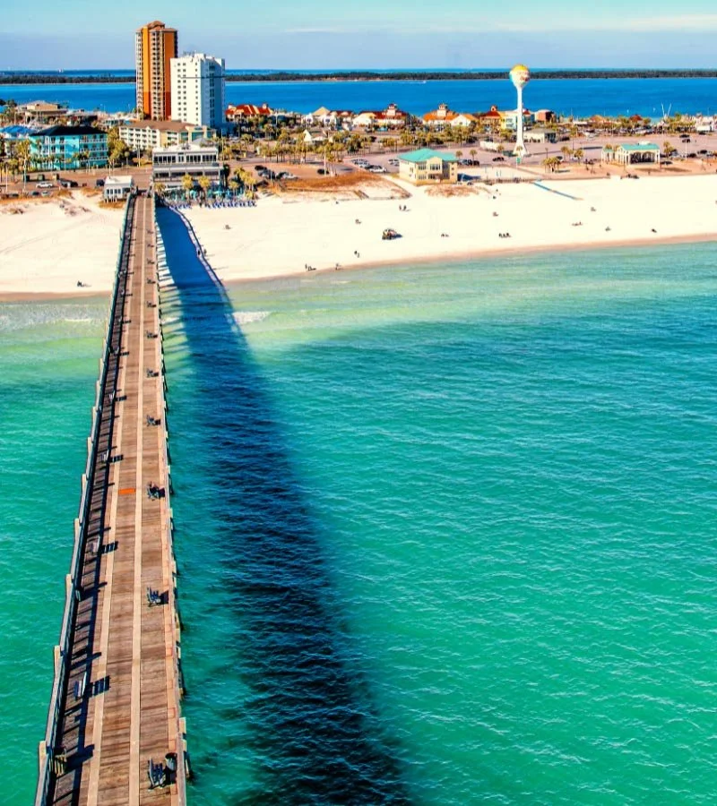 Aerial view of a pier extending into clear turquoise water, leading to a sandy beach with buildings, hotels, and a water tower in the background, under a sunny sky.