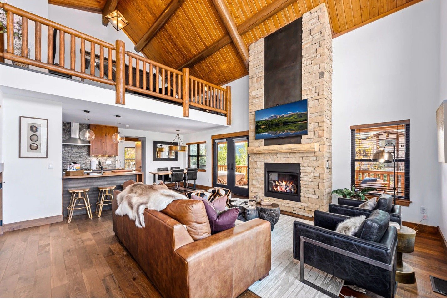 Interior of a spacious living room with a vaulted wooden ceiling, a stone fireplace with a TV mounted above it, leather and fabric sofas, and a view of the kitchen and dining area in the background.