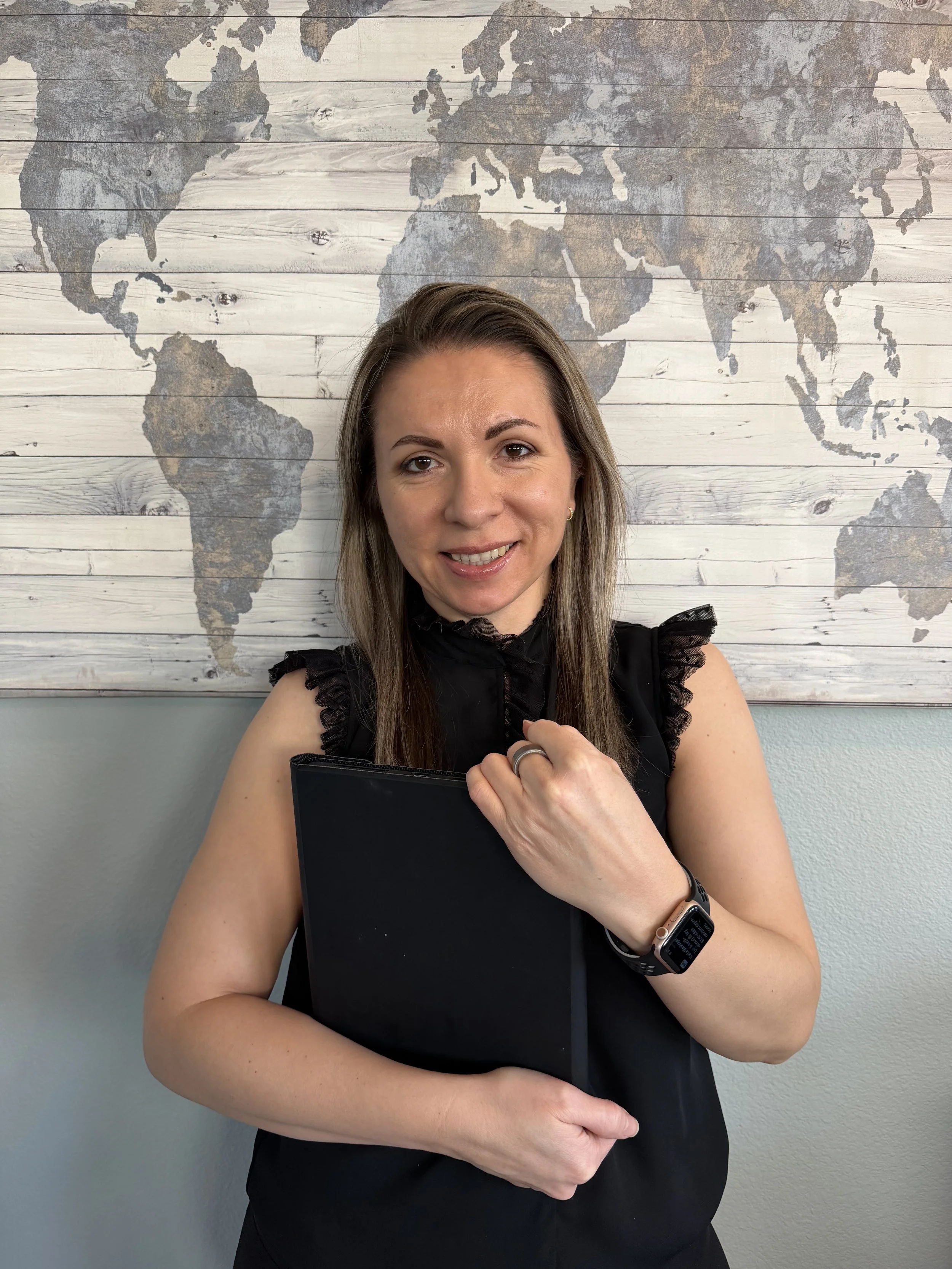 A woman smiling while holding a black folder, wearing a smartwatch, in front of a world map wall decoration.