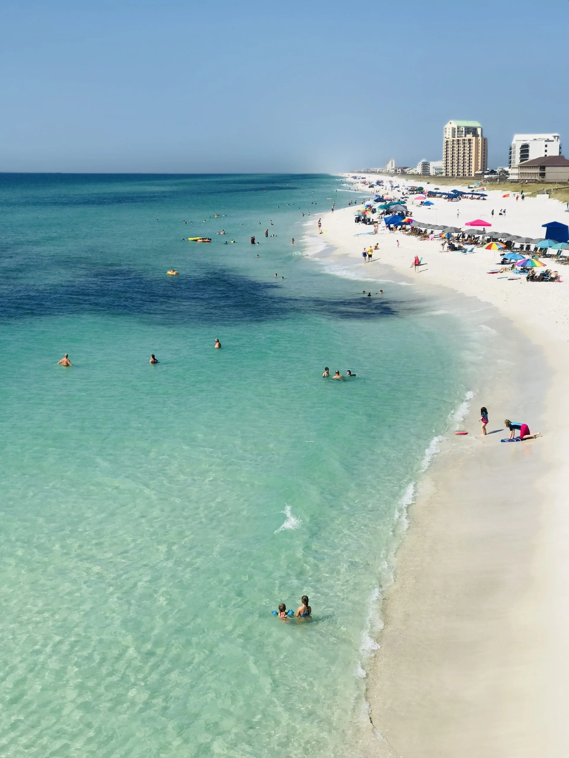 Beach scene with turquoise water, white sand, people swimming and lounging, colorful umbrellas, and high-rise buildings in the background.