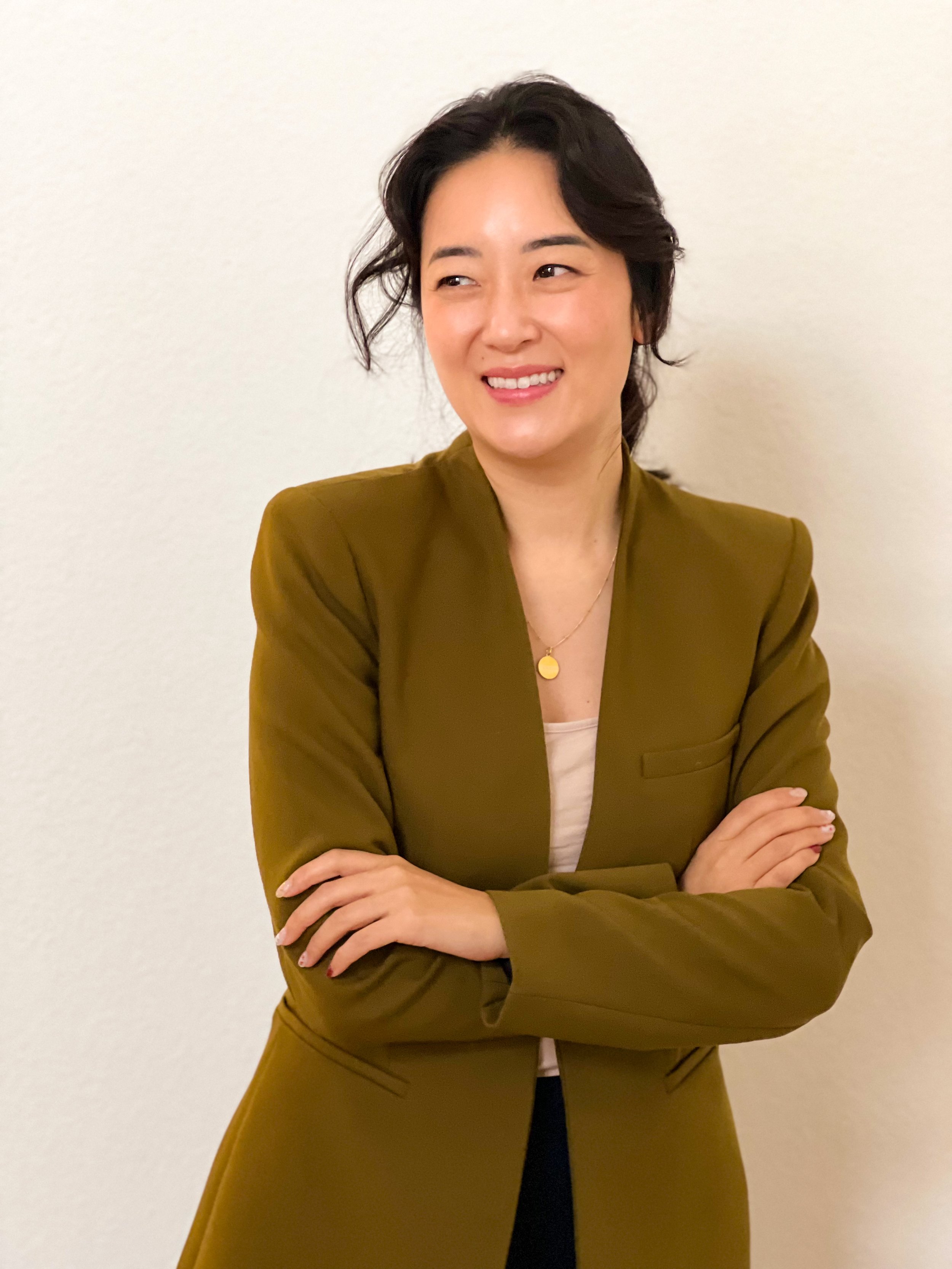 A woman with short black hair, smiling and looking to her right, wearing a brown blazer and a necklace with a gold pendant, standing against a plain off-white wall.