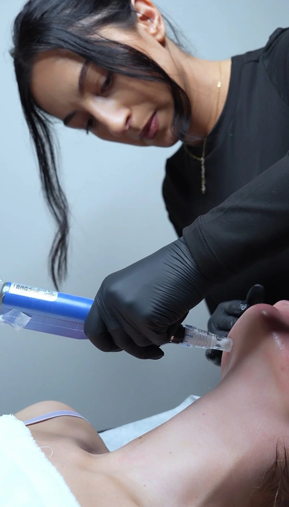 A woman with dark hair and gold jewelry giving a cosmetic injection to a person's chin, wearing black gloves.