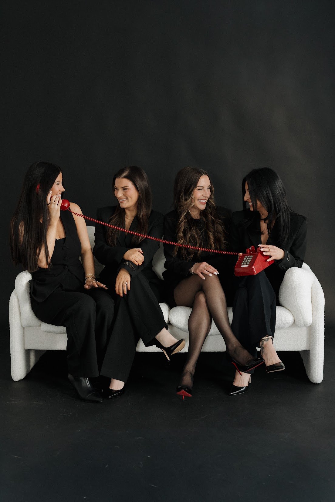 Four women in black outfits sitting on a white sofa against a dark background, engaging in a conversation with one woman holding a red vintage telephone and another woman using a red rotary phone.