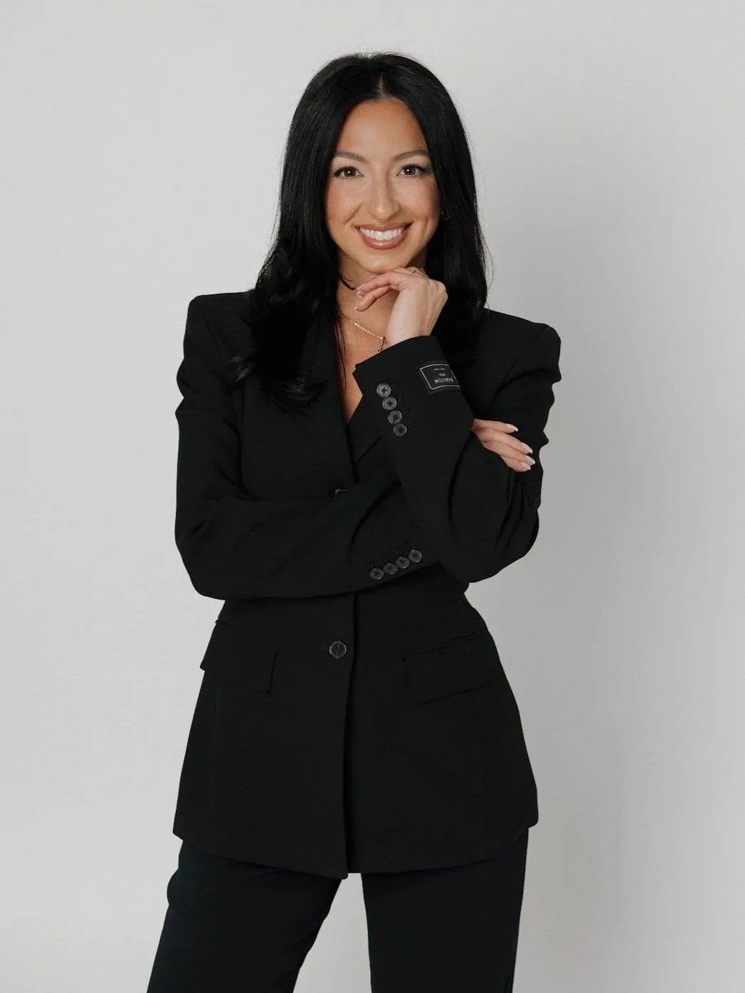 A smiling woman with brown hair in a black top and black pants, holding syringes and wearing gloves, standing in front of a mirror in a modern, well-lit clinic or aesthetic office.