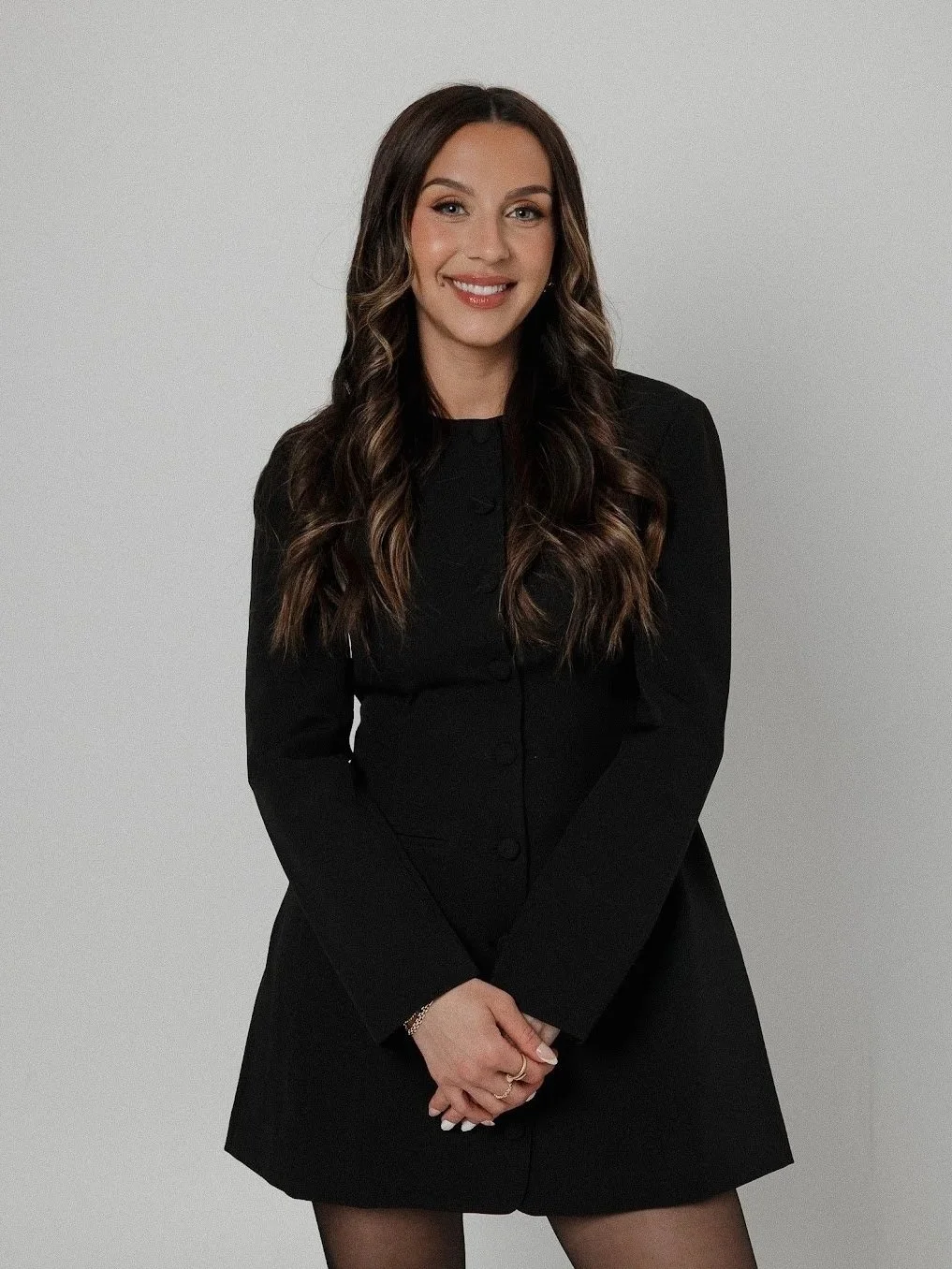 A smiling woman with long, wavy brown hair, wearing a black medical uniform, standing with arms crossed in front of a mirror with a yellow frame, in a medical or office setting.