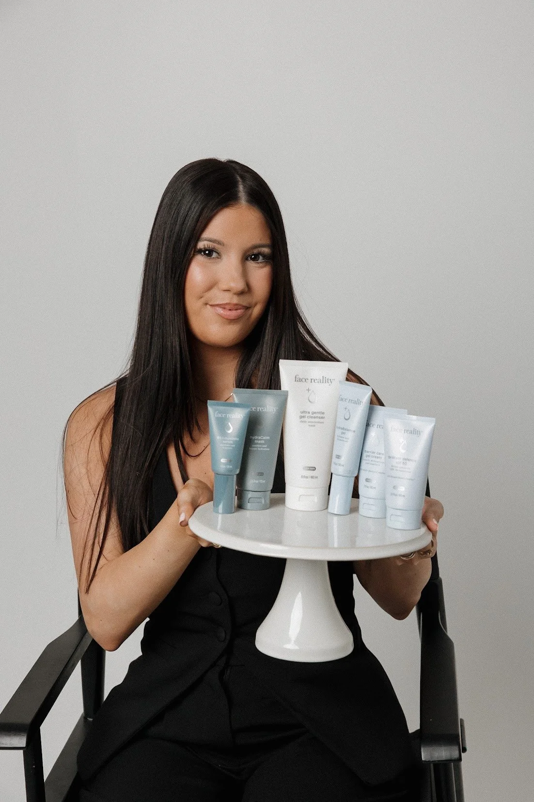 A woman with long dark hair sitting in a black chair, holding a white cake stand with skincare products. The products are labeled Face Reality, and include gel cleansers and other skincare items.