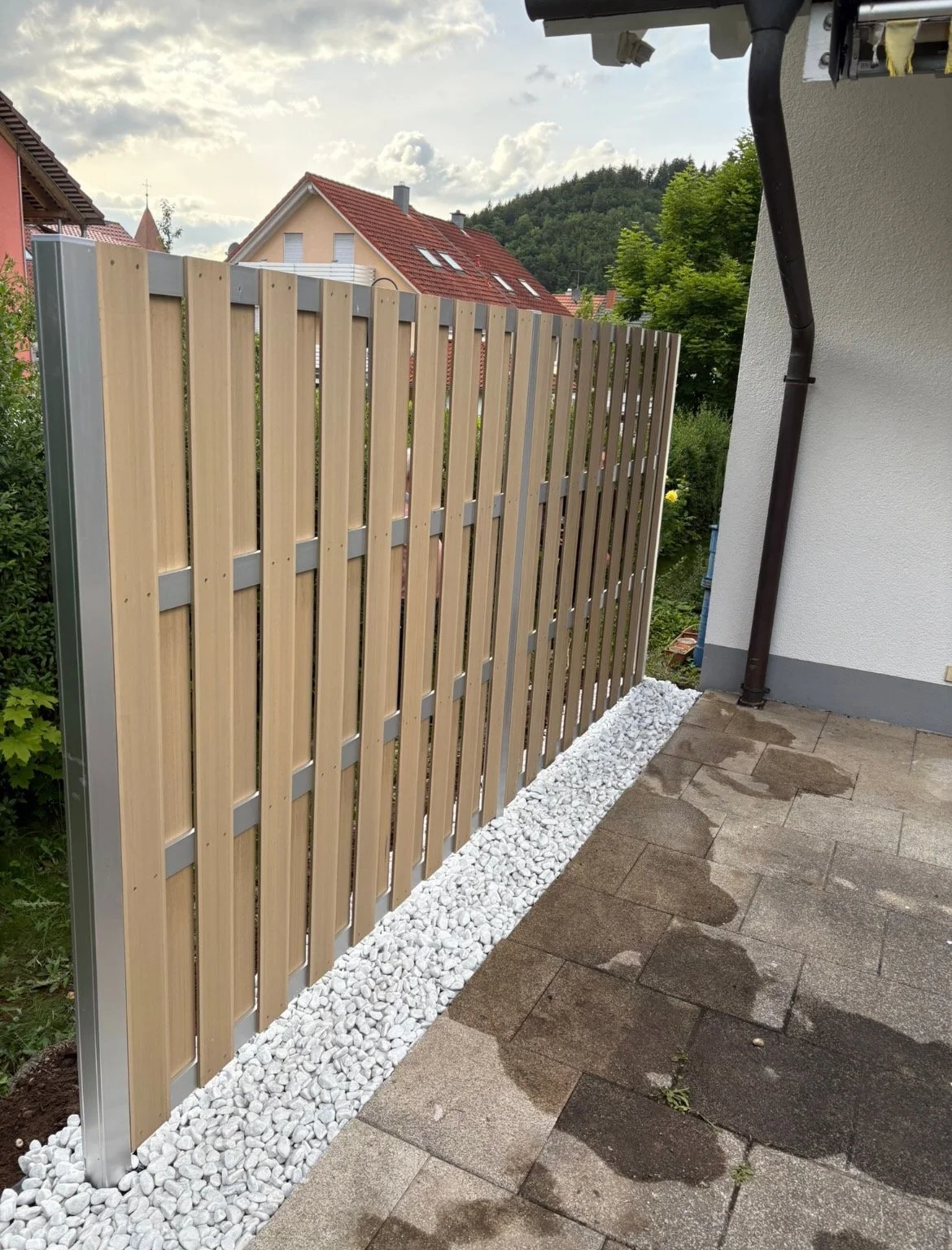 Holzzaun und Pflaster vor einem Haus im Garten, mit Wolken am Himmel.