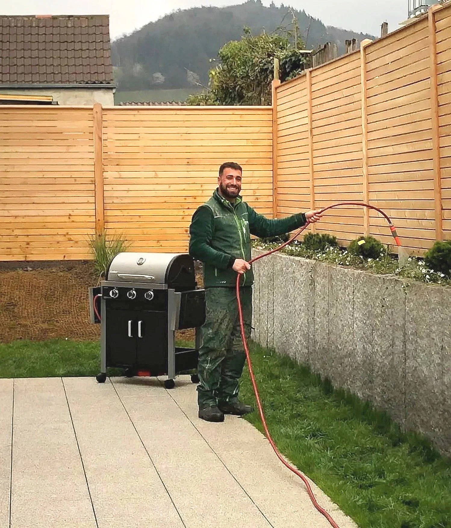 Ein lächelnder Mann beim Gartengrill steht neben einem Grill und gießt Wasser auf den Rasen in einem Garten, im Hintergrund ein Holzlattenzaun und Berge.