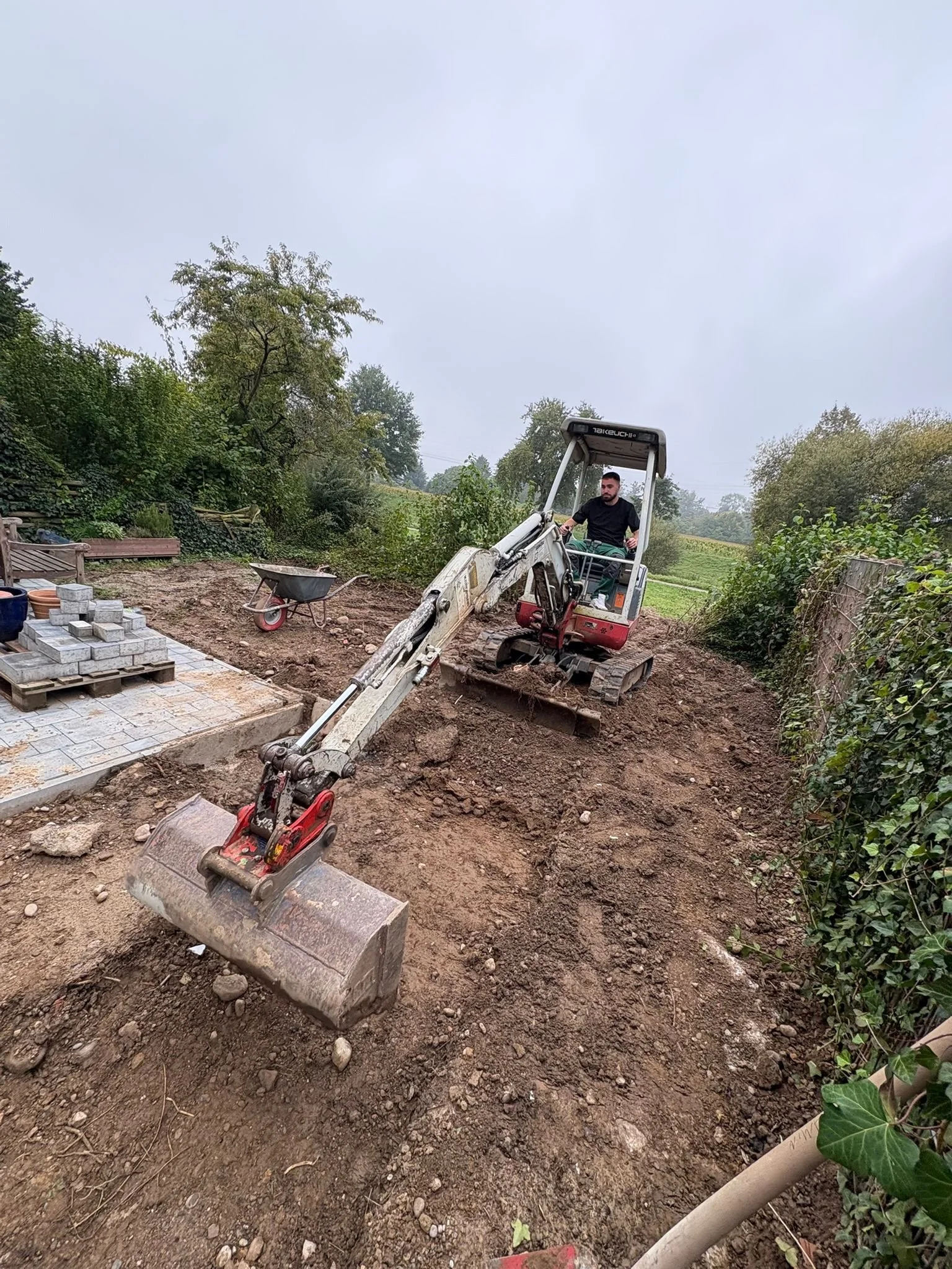 Mann fährt mit einer kleinen Baggermaschine auf einem Garten- oder Baugrundstück, das für eine Pflasterarbeit vorbereitet wird, mit Steinen und Erde umgeben.