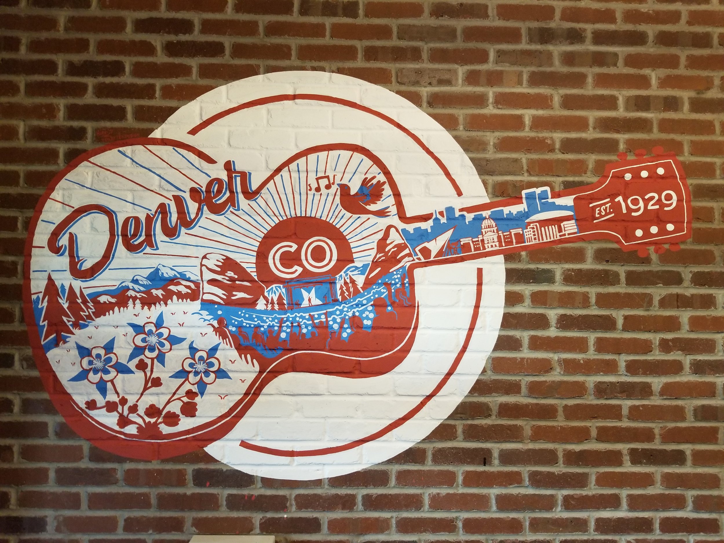Wall art depicting a guitar with illustrations of mountains, flowers, and cityscape, featuring the words 'Denuet Co' and 'Est. 1929' on the headstock, with a background of a brick wall.