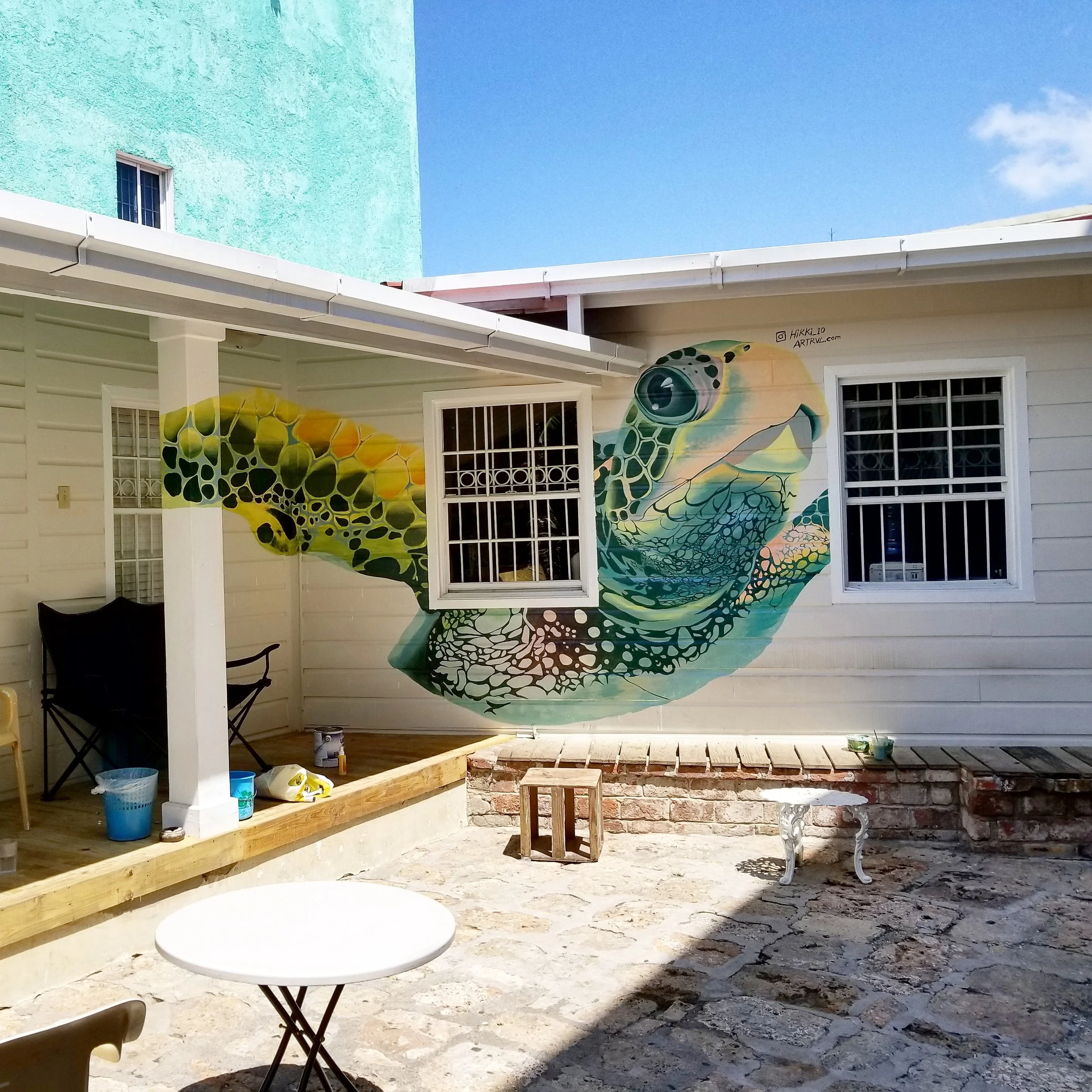 Colorful mural of a sea turtle painted on the side of a white house with barred windows and a small porch, under a bright blue sky.