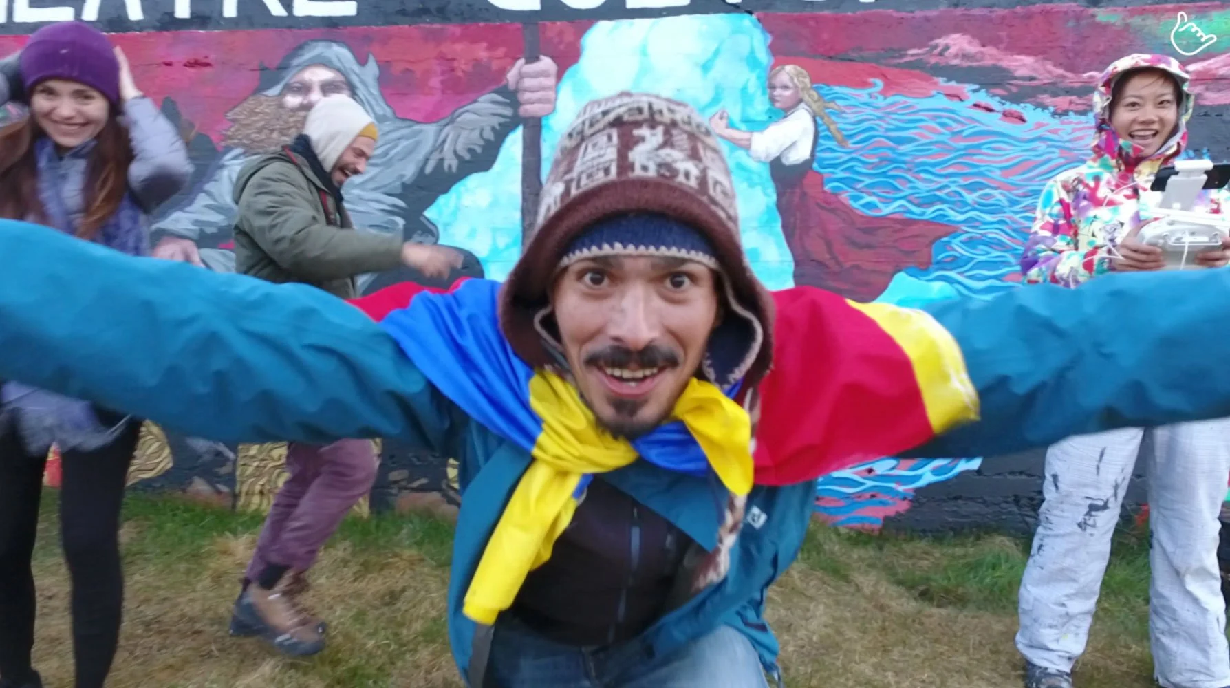 Group of five people outdoors in front of a colorful mural, smiling and posing for the camera, wearing rain jackets and hats.