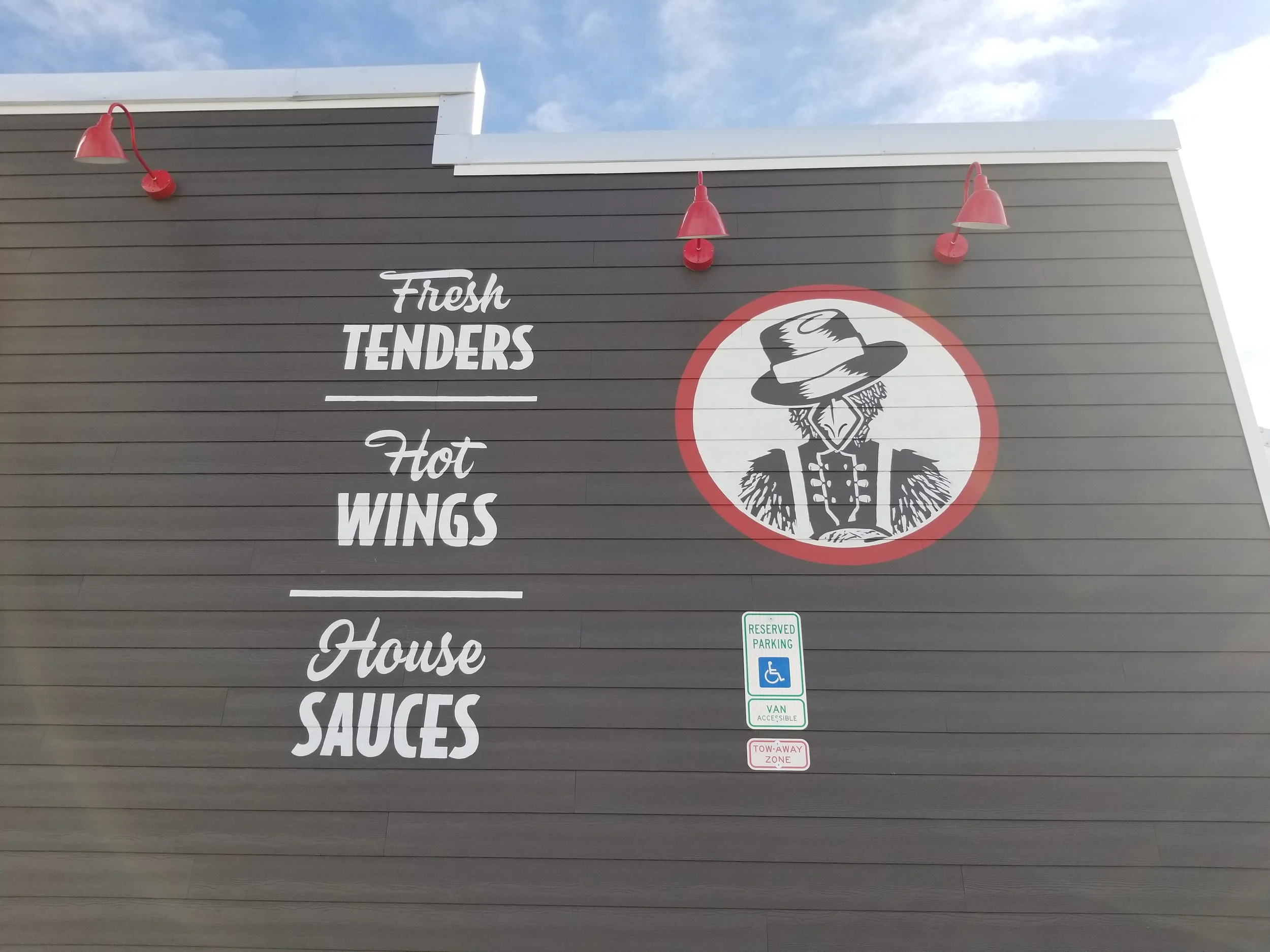 Restaurant exterior wall with sign listing menu items: fresh tenders, hot wings, house sauces, and a graphic of a scarecrow wearing a hat and overalls.
