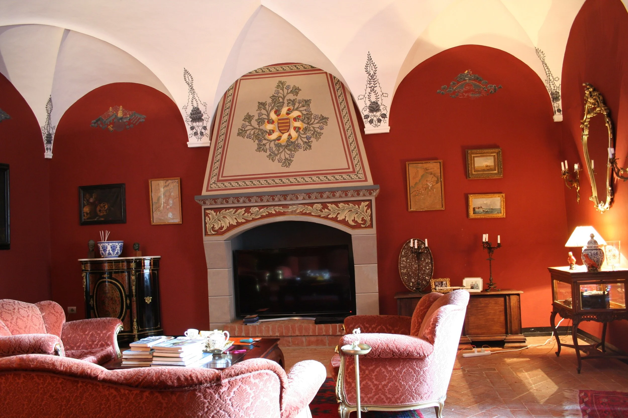 A vintage living room with red walls, ornate decorations, floral-patterned pink sofas, a wooden coffee table with books, a television set in front of a fireplace, and various antique artworks and lighting fixtures.