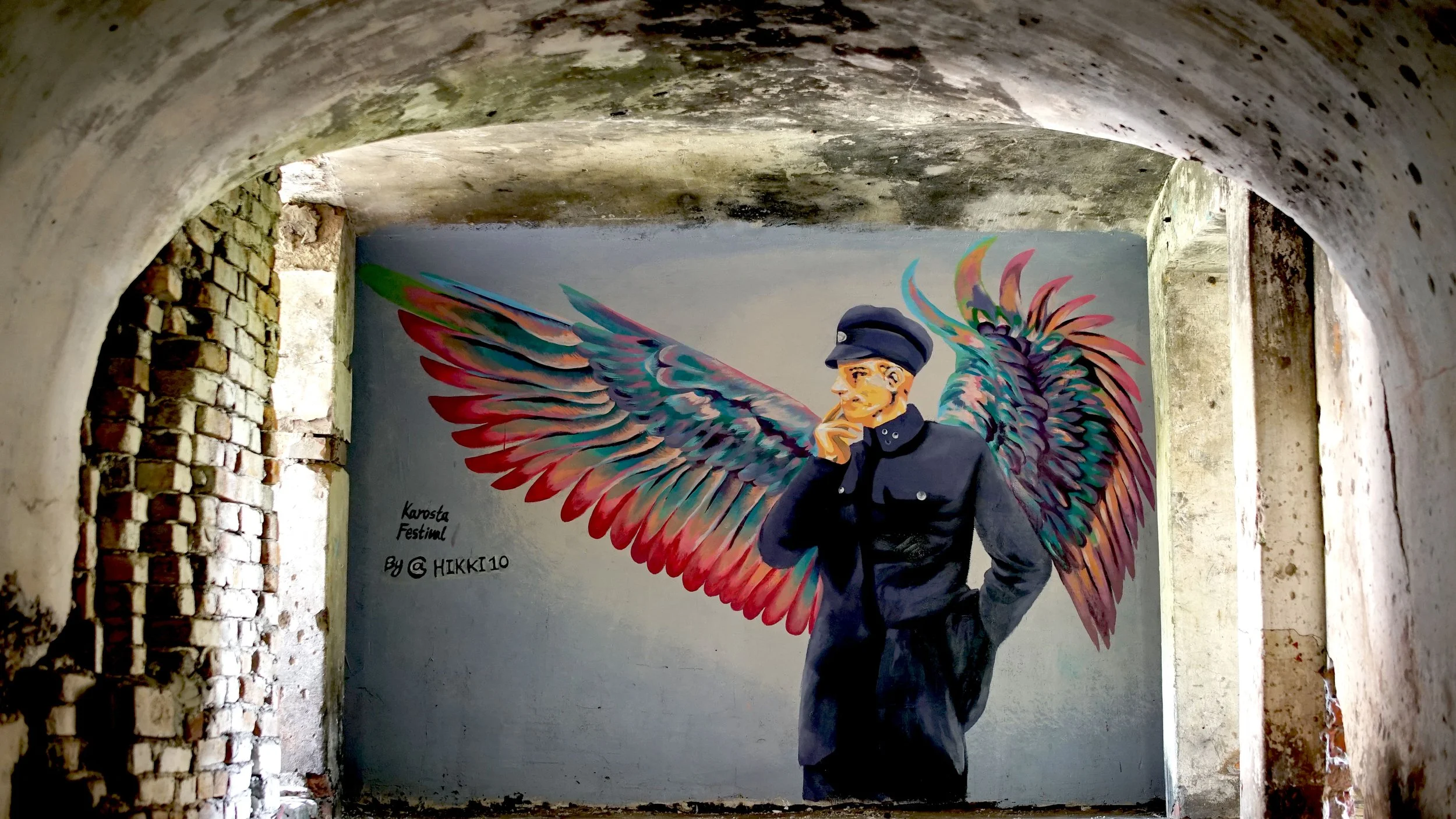 Street art mural of a man in a military uniform with multicolored wings on his back, under an archway with exposed brick and concrete surfaces.