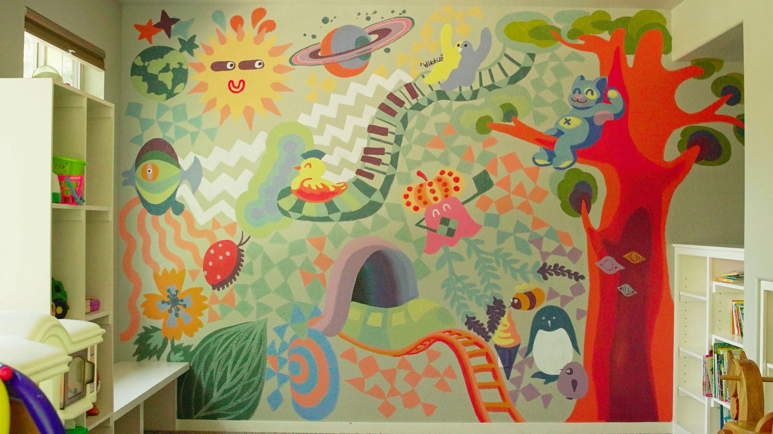 Colorful mural featuring a smiling sun with sunglasses, a whale, a duckling, a rainbow, a planet with rings, animals like a cat and penguin, and various abstract shapes and patterns in a child's playroom.