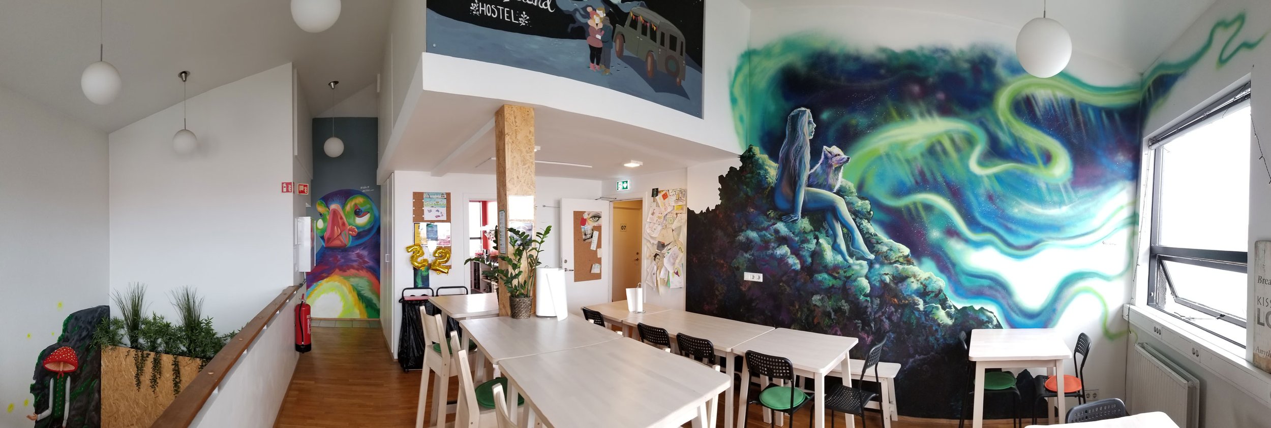 Interior of a room with vibrant colorful murals, including a mythical creature on rocks and a portrait with swirling green and blue patterns, white tables and chairs, and windows letting in natural light.