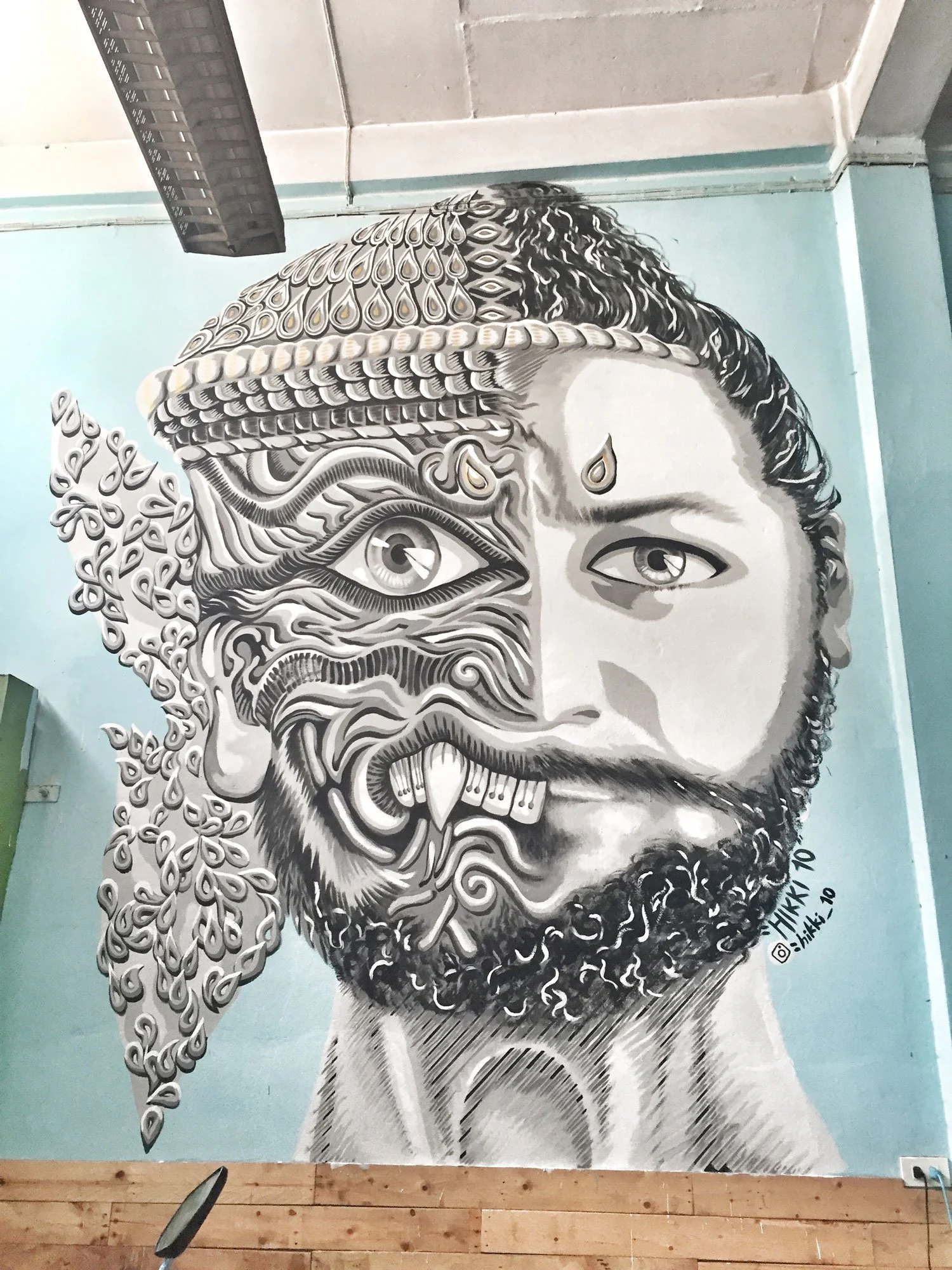 Street art mural of a person's face split into two halves, combining realistic and abstract elements, with intricate black and white patterns, a beard, and various stylized facial features, created in a contemporary graffiti style.