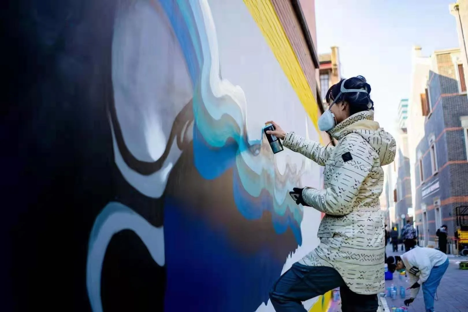 A person wearing a face mask, gloves, and a patterned jacket spray paints a mural on a large outdoor wall in an urban area. The mural features abstract, colorful swirls.