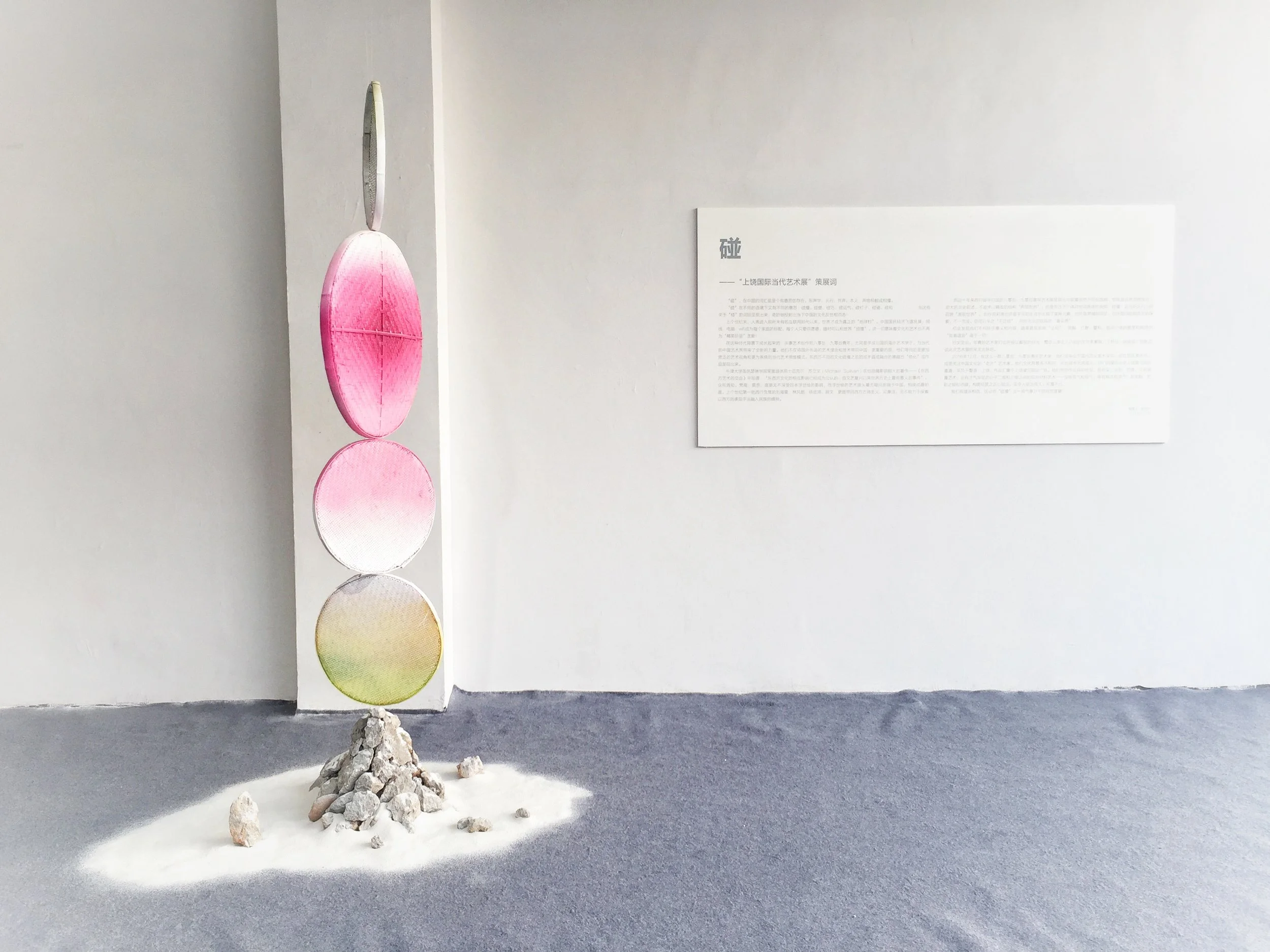 Art installation with four large, vertically aligned circular panels in pink, white, and green, mounted on a white vertical structure, with a pile of rocks and white powder at the base, displayed in an art gallery with a white wall and informational 