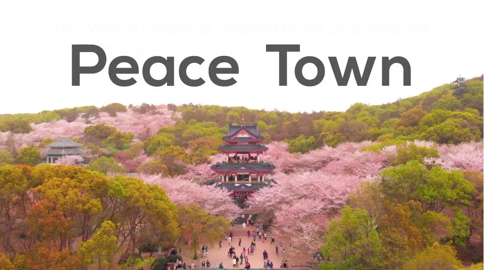 A scenic view of a traditional Asian pagoda surrounded by pink cherry blossom trees and green hills with people walking in the foreground. The words 'Peace Town' are written above in large gray letters.