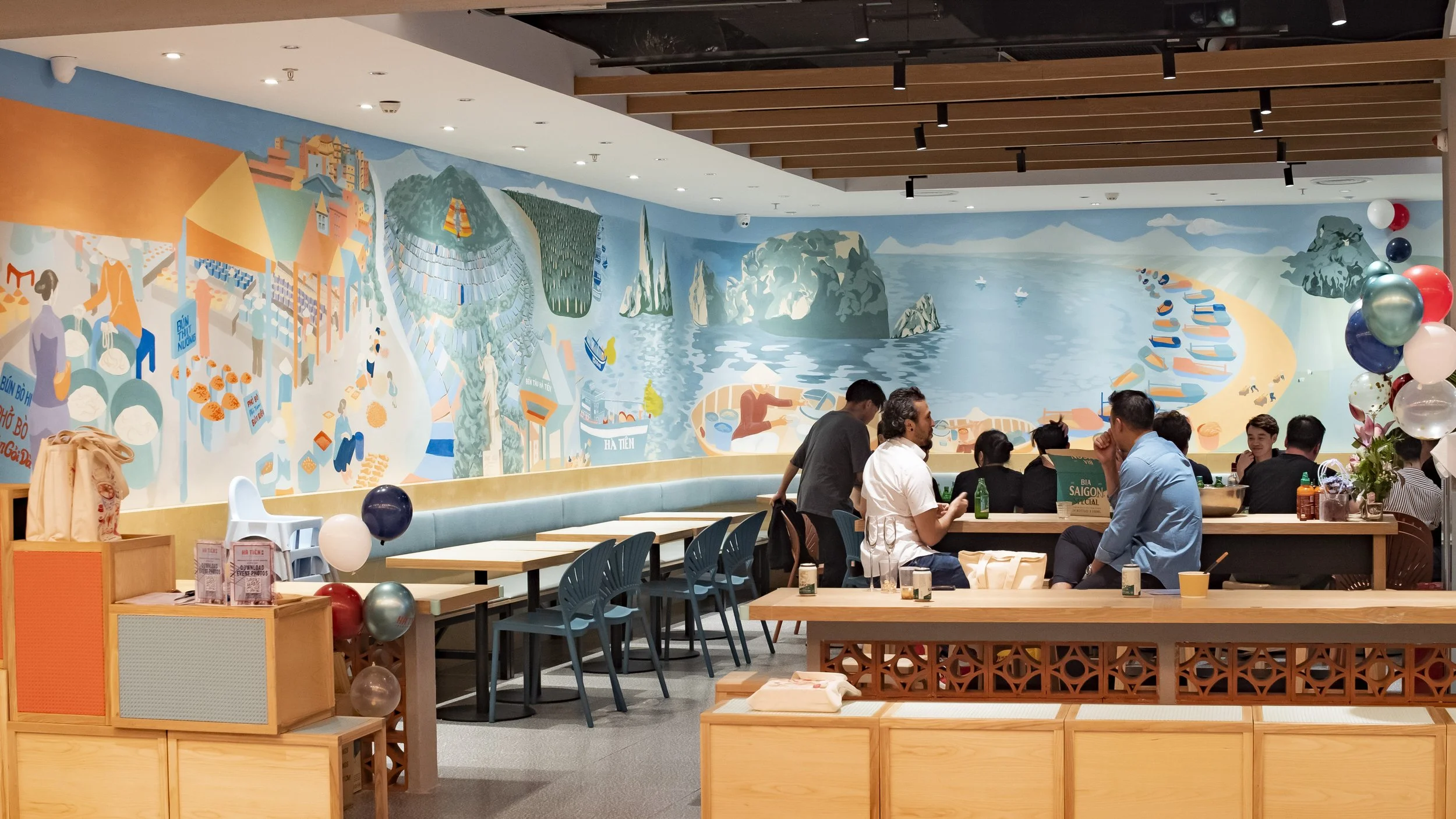 A restaurant interior with a large colorful mural of a coastal scene on the wall, including mountains, water, boats, and people. There are tables, chairs, and a group of people sitting and talking.