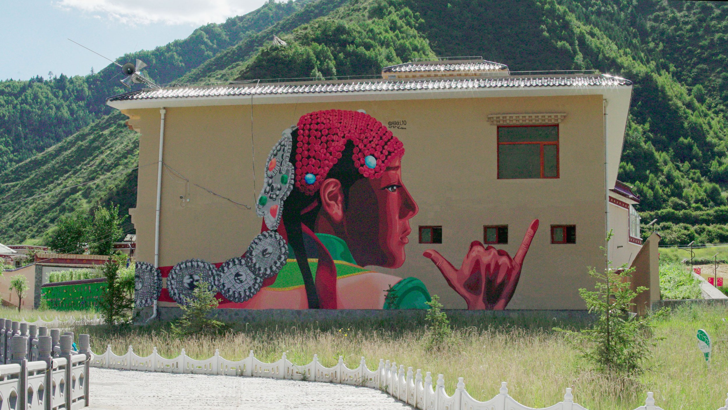 Large mural of a woman in traditional attire with pink hair and jewelry, making a gesture with her hand, painted on the side of a building in a mountainous area.