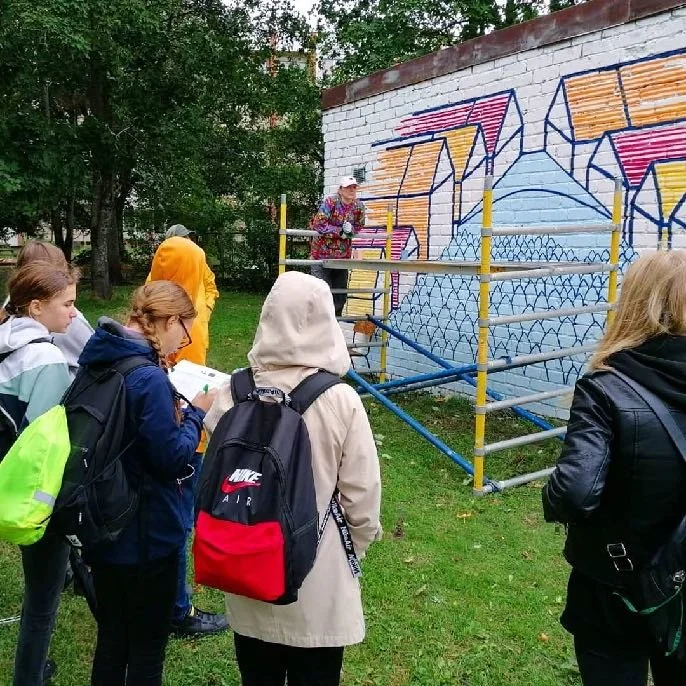 Group of young people gathered outdoors, observing a woman standing on scaffolding painting graffiti on a white brick wall.