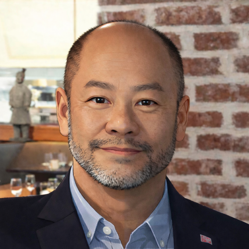 A middle-aged Asian man with a beard, wearing a light blue collared shirt and black blazer, smiling in a restaurant with a brick wall and a decorative sculpture in the background.