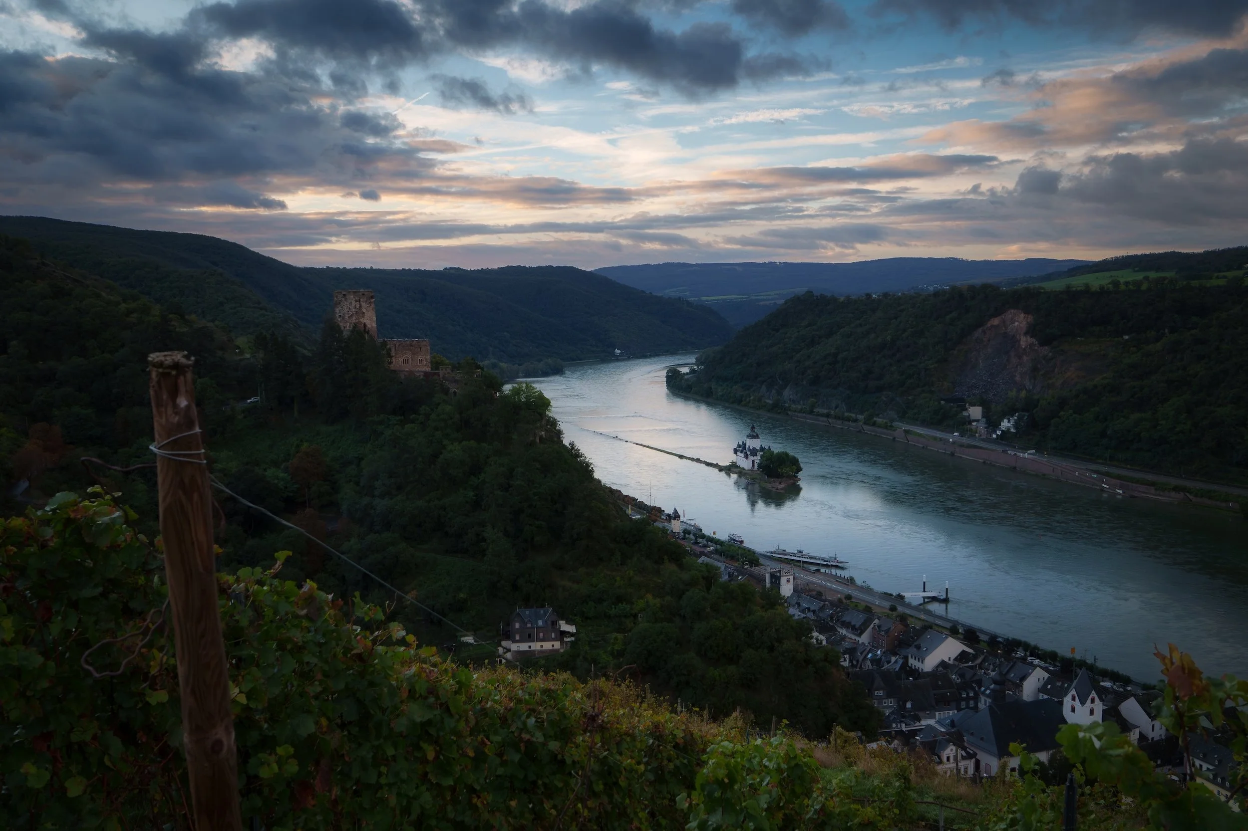 Blick auf einen Fluss, umgeben von Hügeln mit einem kleinen Dorf am Ufer und einem Schloss auf einem Hügel, bei Sonnenuntergang oder Dämmerung, bewölkter Himmel.