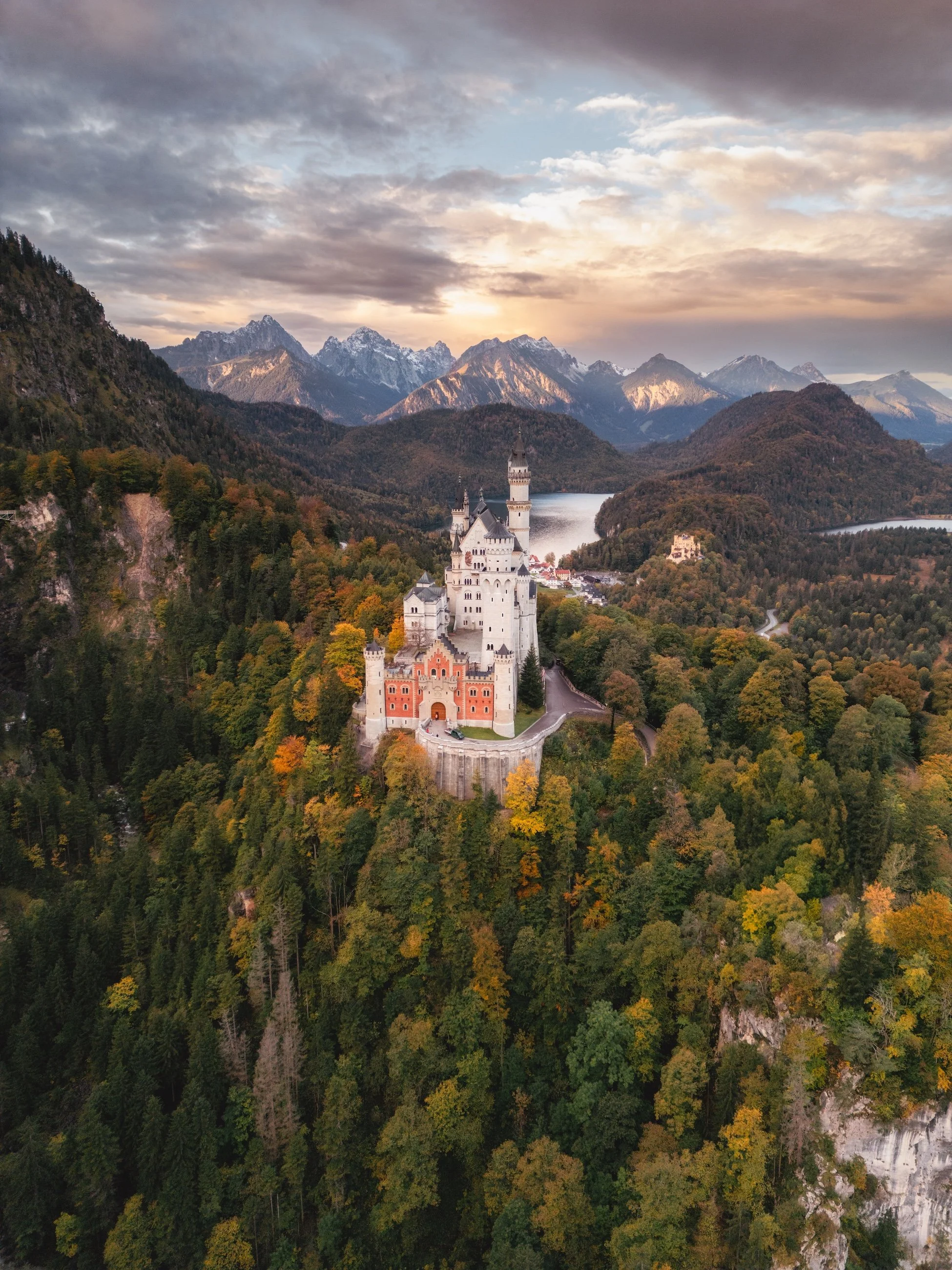 Burg auf einem Hügel, umgeben von dichtem Herbstwald, mit Bergen im Hintergrund bei Sonnenuntergang.