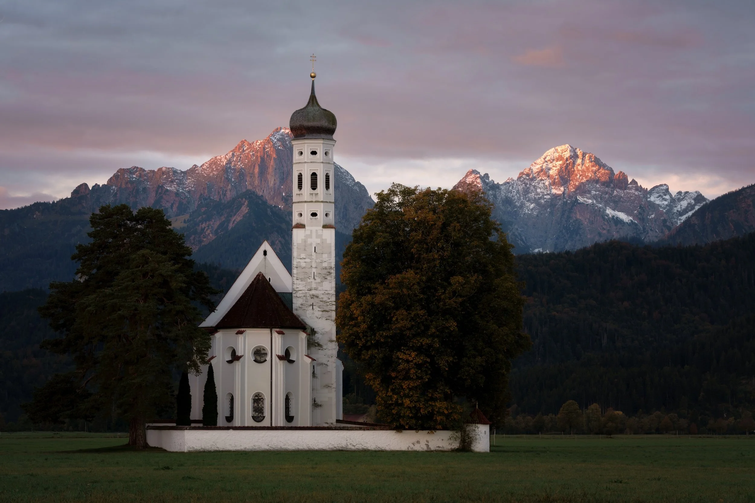 Eine weiße Kirche mit dunklem Turm und steilen Dächern steht auf einer grünen Wiese, im Hintergrund erheben sich schneebedeckte Berge bei Sonnenuntergang.