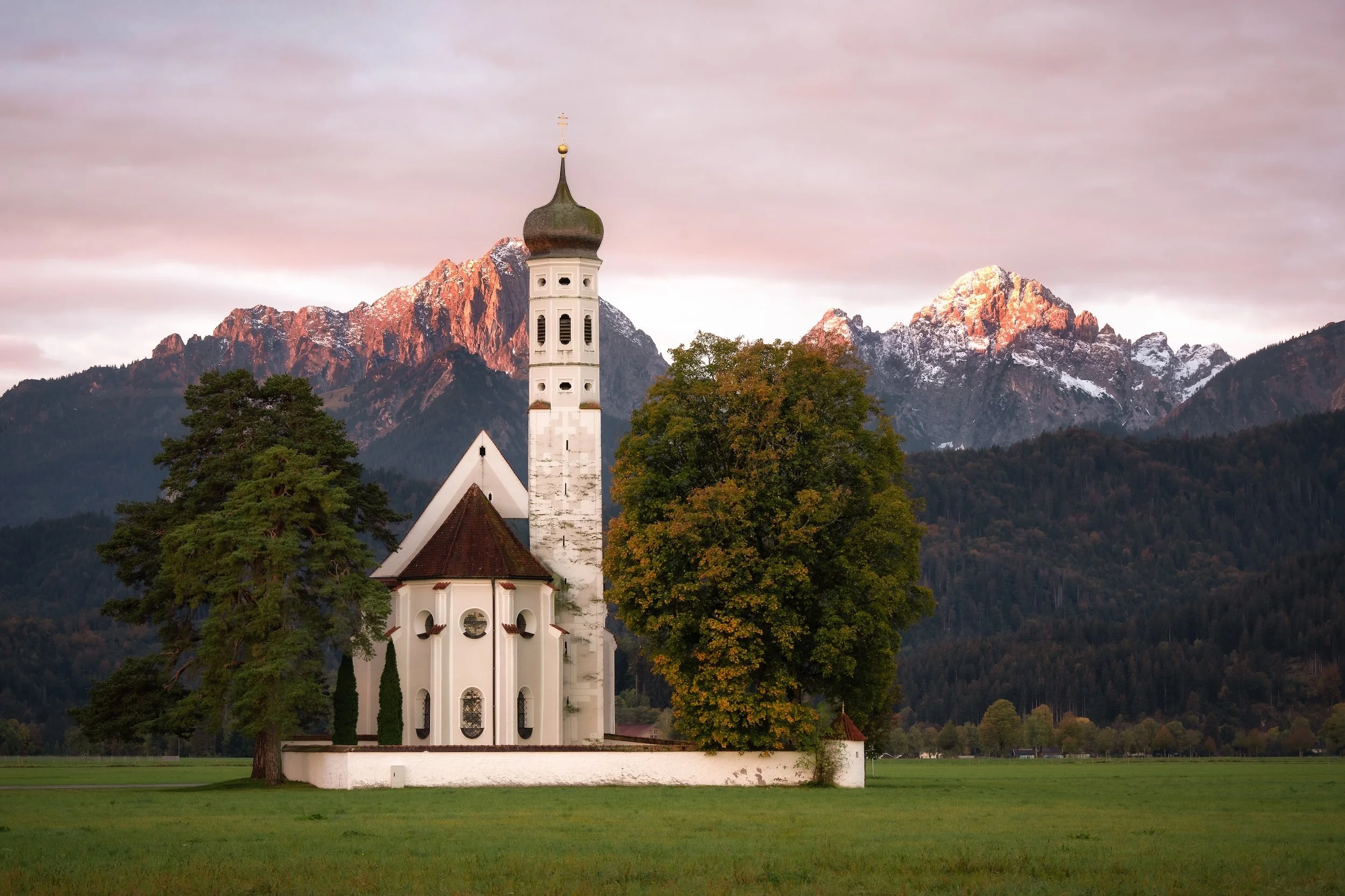 Eine weiße Kirche mit dunklem Dach und Turm, umgeben von Bäumen, mit Bergen im Hintergrund bei Sonnenuntergang.