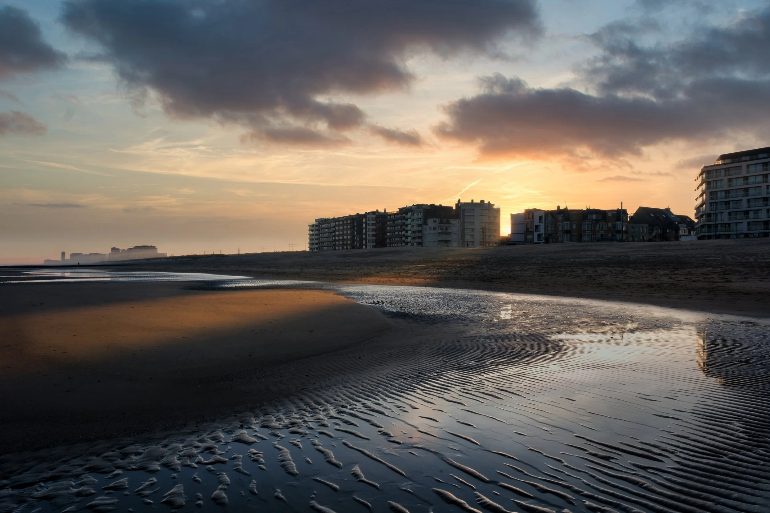 Strand bei Sonnenuntergang mit Gebäuden im Hintergrund, gespiegelt im Wasser mit Sandmustern im Vordergrund