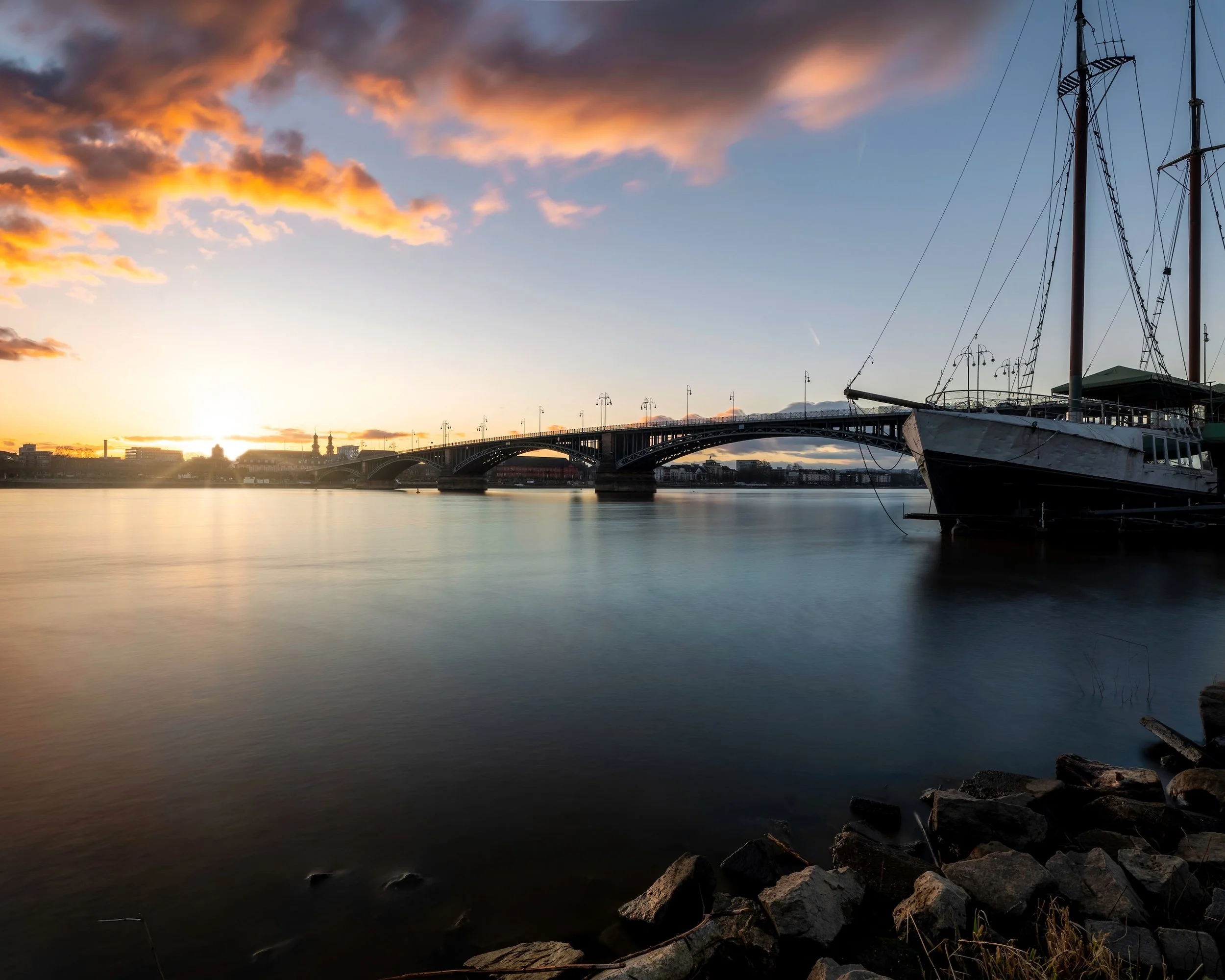 Ein Sonnenuntergang über einer Brücke und einem Fluss mit einem anlegenden Segelschiff im Vordergrund.