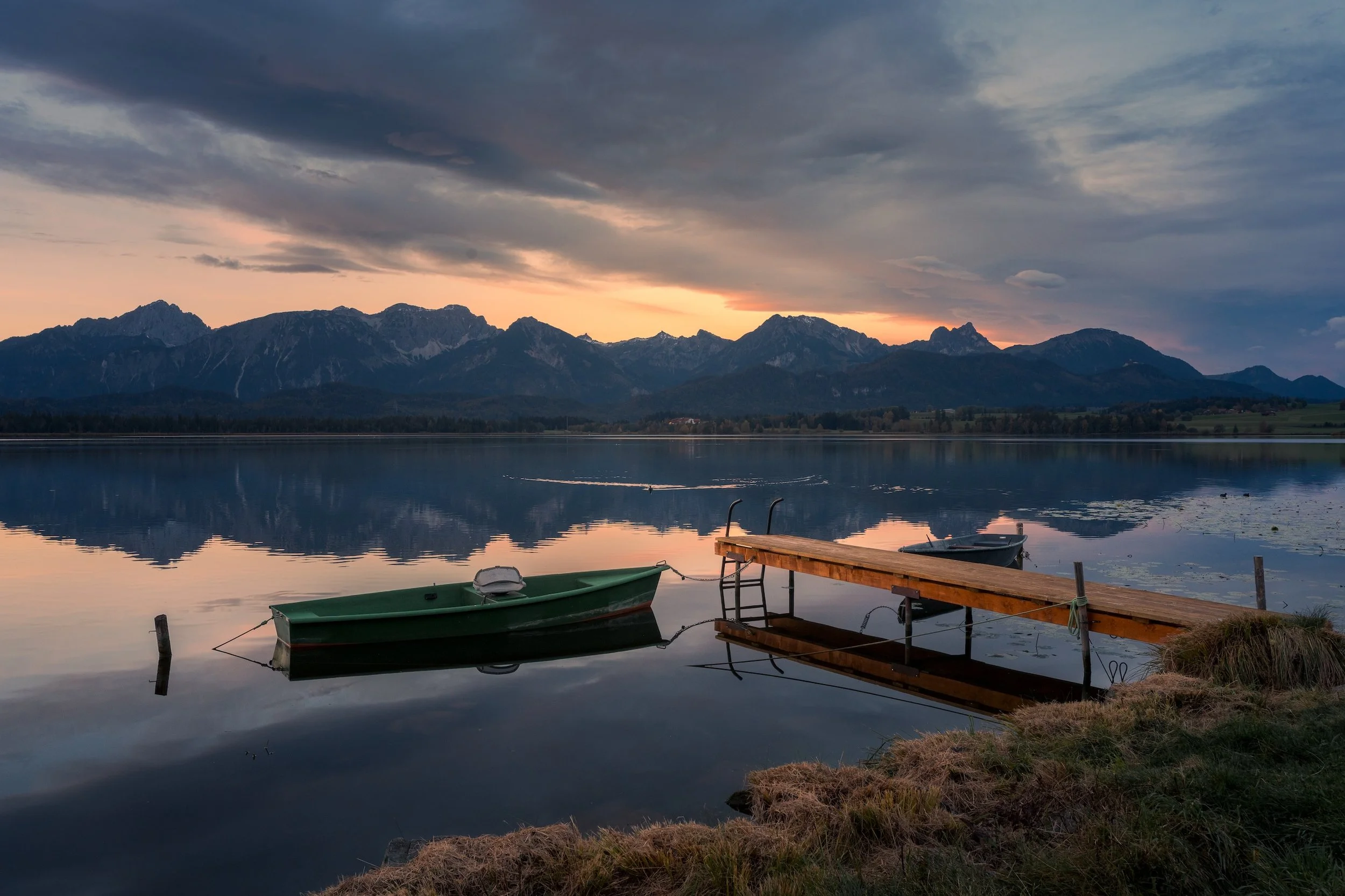Ein ruhiger See mit einem kleinen Steg und zwei Booten bei Sonnenuntergang, im Hintergrund Berge und bewölkter Himmel.