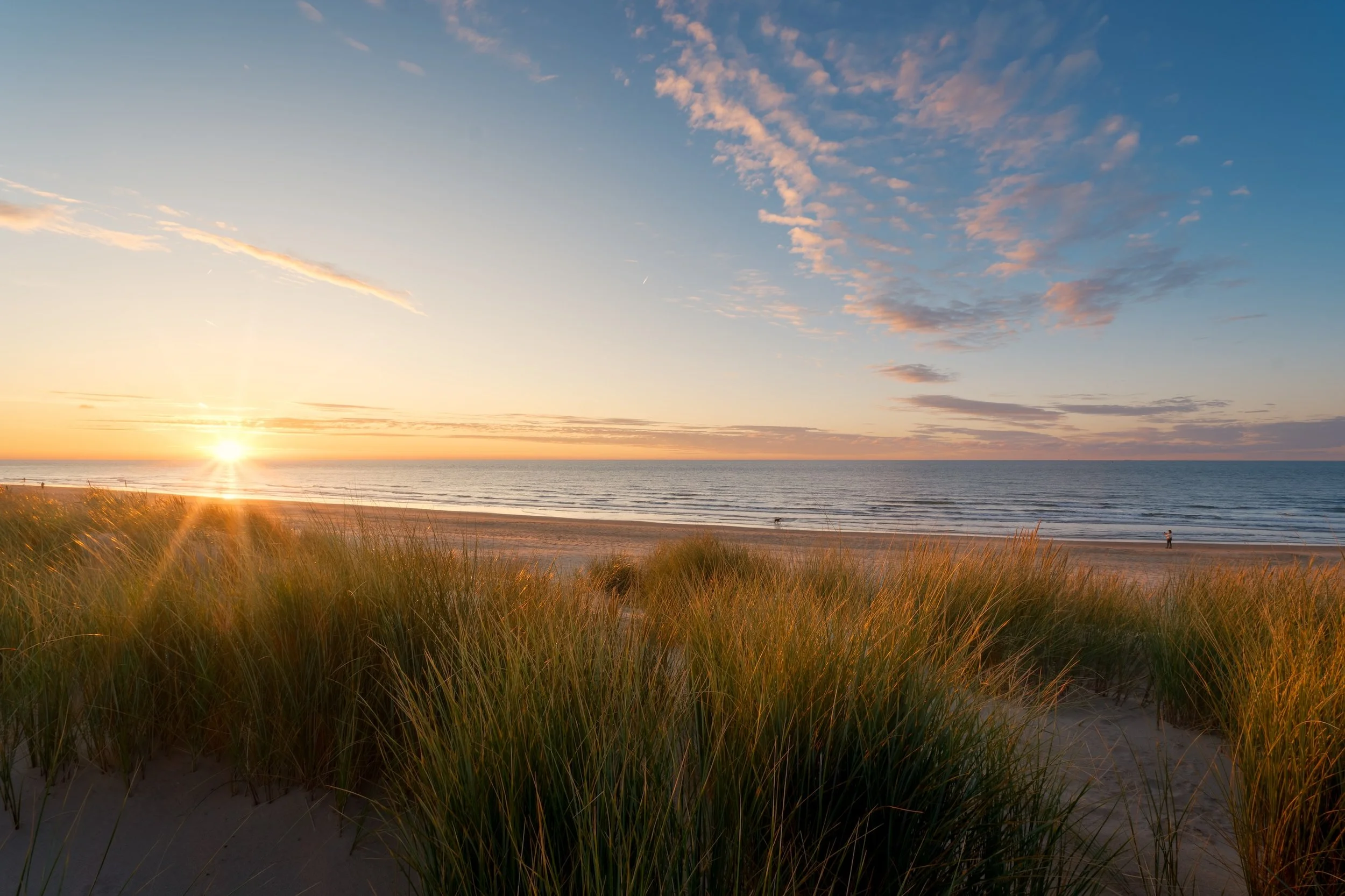 Ein Sonnenuntergang über einem Sandstrand mit Gras und dem Meer im Hintergrund, einige Menschen sind am Wasser zu sehen.